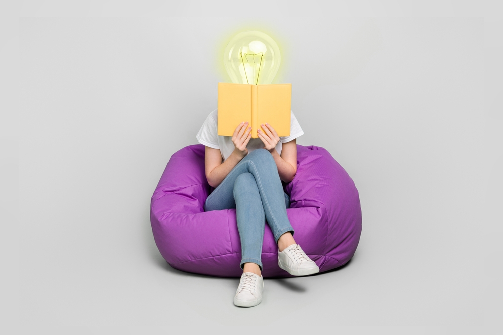 A woman with a bright electric bulb as a head reading a book sitting on a purple beanbag with grey background